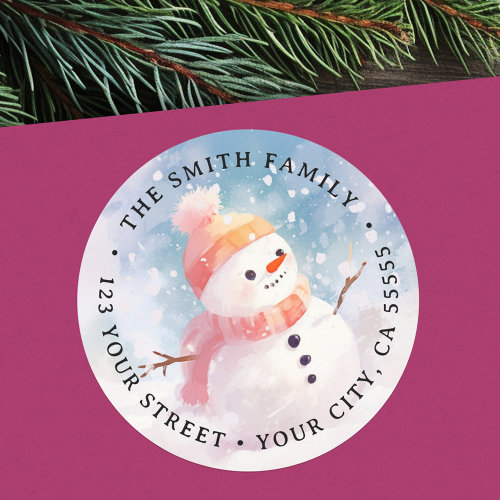 Round watercolor snowman return address label featuring a cheerful snowman with a peach hat, pom-pom, and scarf on a snowy blue background, customizable with name and address. Perfect for Christmas cards, holiday invitations, and festive seasonal mail.